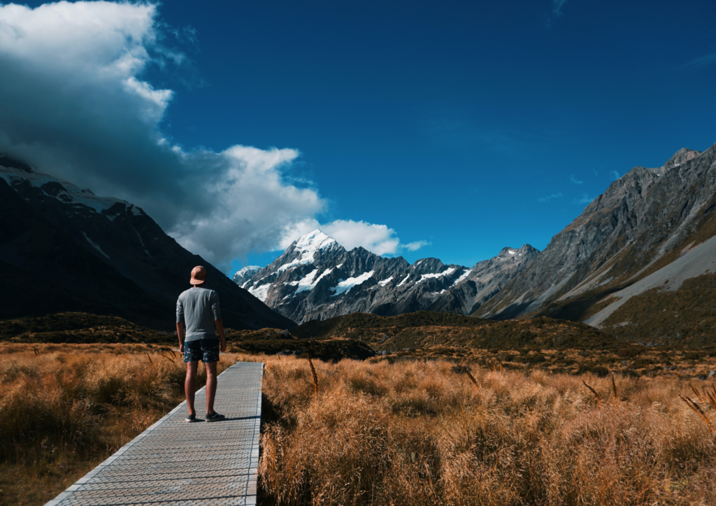 Young person, possibly an engineer or infrastructure asset manager, standing on a boardwalk look at Mount Cook in Canterbury, New Zealand, Aotearoa, which is a snow covered mountain. He's also surrounded by what looks like tussock grass. It's a sunny day, with blue skies and a bit of cloud.